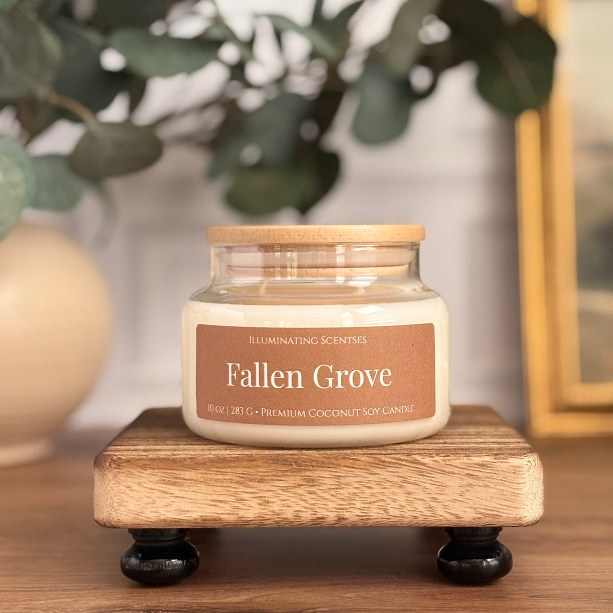 A jar candle labeled 'Fallen Grove' by Illuminating Scents sits on a small wooden stand on a wooden table, with blurred green leaves and a framed picture in the background.