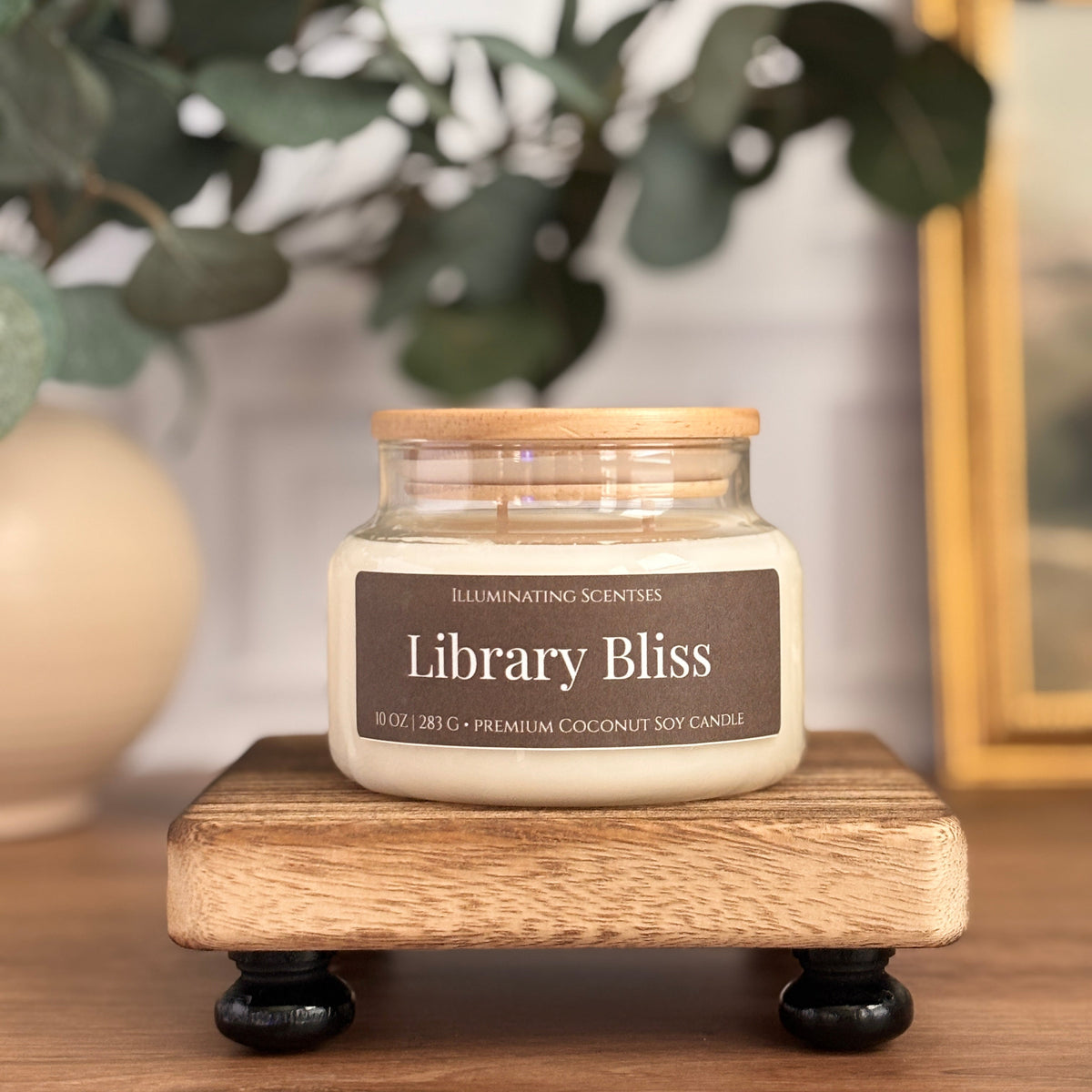 A jar candle labeled 'Library Bliss' by Illuminating Scents, placed on a small wooden stand on a wooden table with blurred greenery and a framed picture in the background.