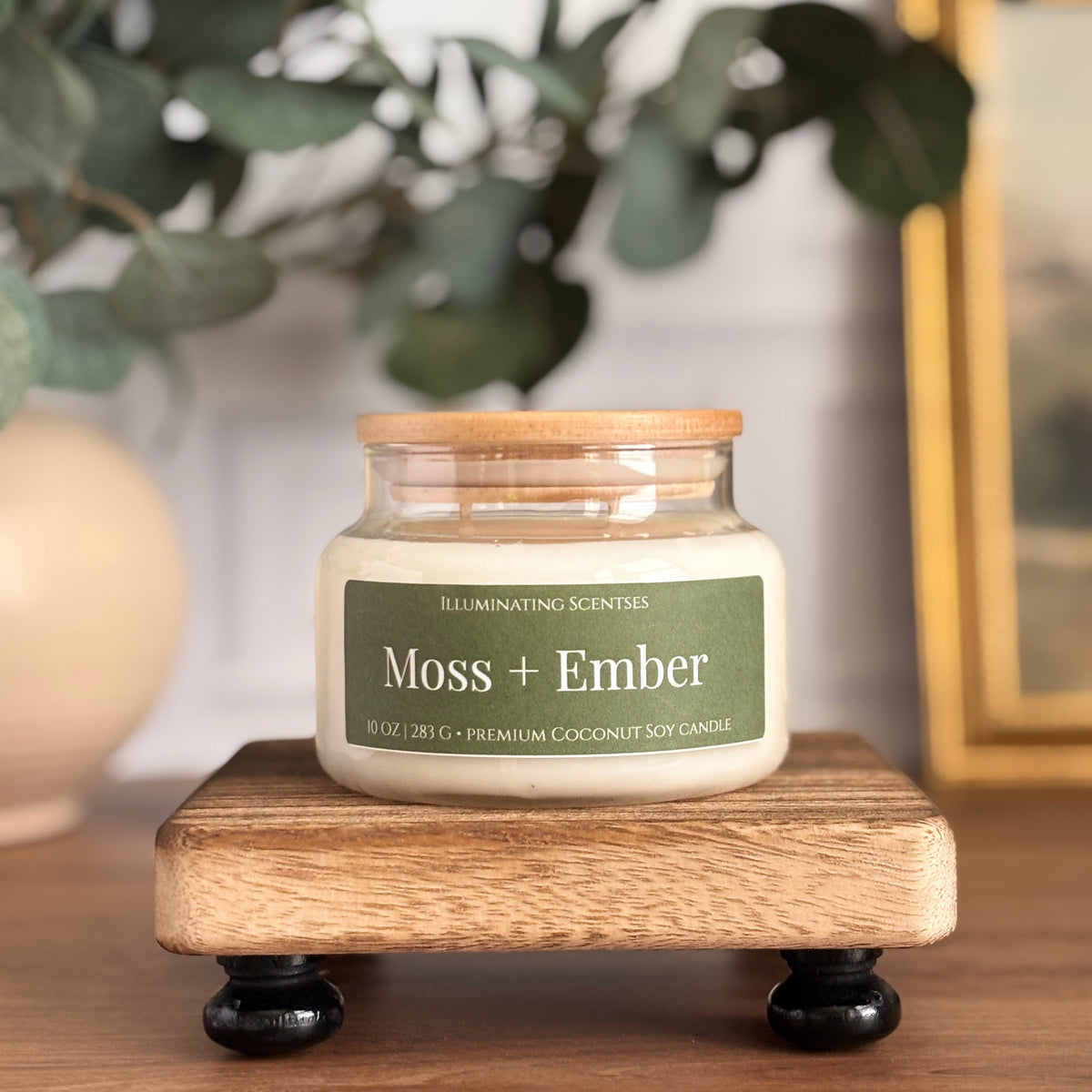 A jar candle labeled 'Moss + Ember' by Illuminating Scentsie sits on a small wooden stand. The candle has a light wood lid and a green label, set on a wooden tabletop with blurred eucalyptus leaves and a framed picture in the background.