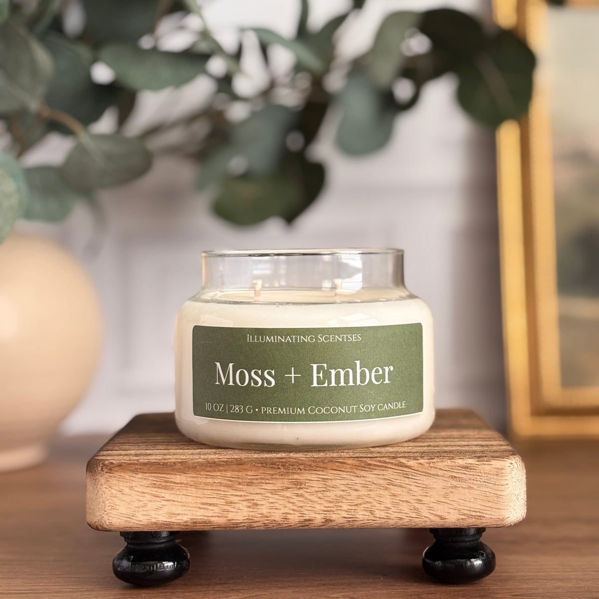 A glass jar candle labeled 'Moss + Ember' by Illuminating Scents sits on a small wooden stand with black feet. The candle is styled on a wooden table with blurred green leaves and a gold picture frame in the background.